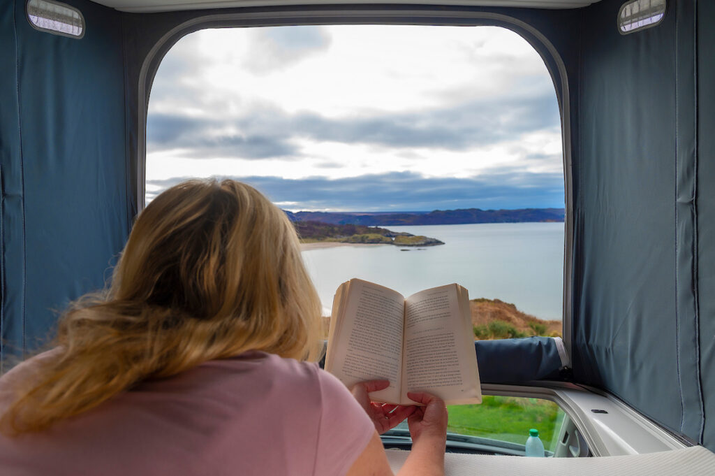 girl reading book in campervan roof overlooking Scottish coastline with Four Seasons Campers campervan rental