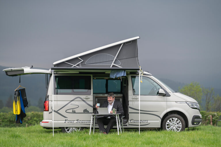 man working beside campervan with wet suit, trunks and flippers and holding up coffee cup with Four Seasons Campers corporate travel Scotland