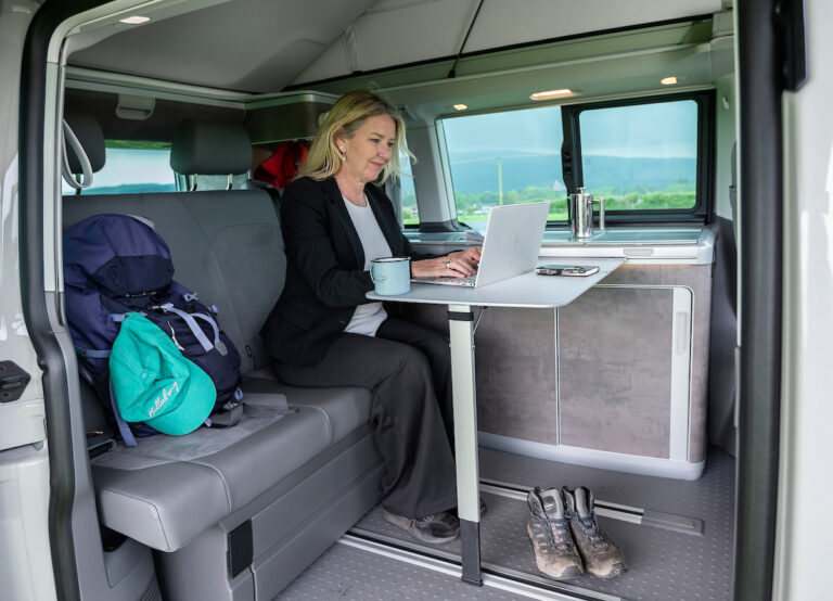 woman in suit working in campervan with walking boots with pop up roof by loch in Scotland for business travel accommodation with Four Seasons Campers